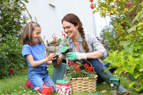 A woman and a young girl are gardening outdoors in a lush, well-maintained garden, sitting on the grass and surrounded by various plants and flowering bushes. The woman, wearing a grey jacket and green gardening gloves, is smiling as she hands a potted red flowering plant to the girl, who is dressed in a blue t-shirt and denim shorts. They are engaged in planting or tending to the garden, with gardening tools and a woven basket nearby. The garden features a mix of vibrant green foliage, blooming flowers, and neatly trimmed hedges, with a white wall and a glimpse of a garden structure in the background. The scene suggests a peaceful, sunny day, capturing a moment of outdoor gardening activity that aligns with professional gardening and landscaping services offered by Hedge Trimming Kentish Town, supporting local gardening maintenance and outdoor care in the area near postcode N7, Kentish Town.