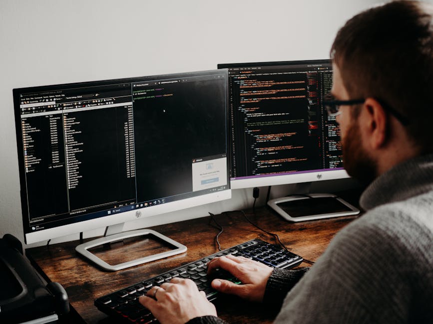A man with short brown hair, glasses, and a beard is working at a wooden desk with two large dual-monitor screens displaying lines of code and software interfaces. His right hand is on a black keyboar